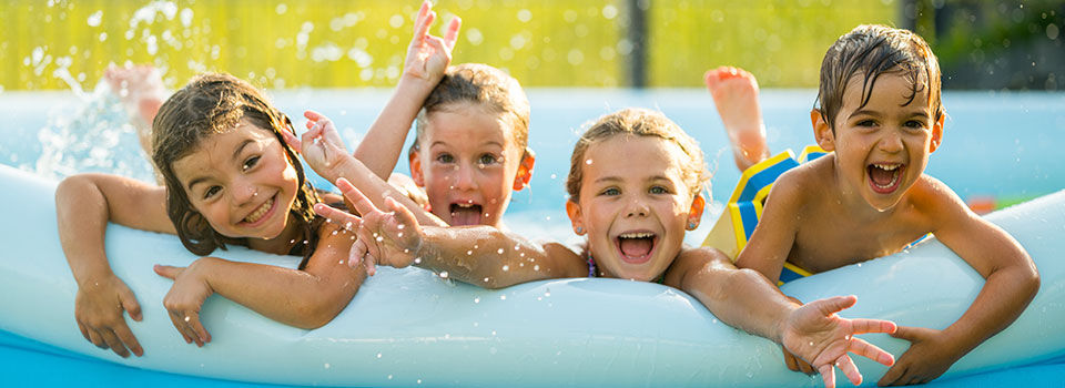 Four children are smiling on the edge of a backyard inflatable pool. The pool is blue and aqua colored, and there is water splashing in the background as they kick.