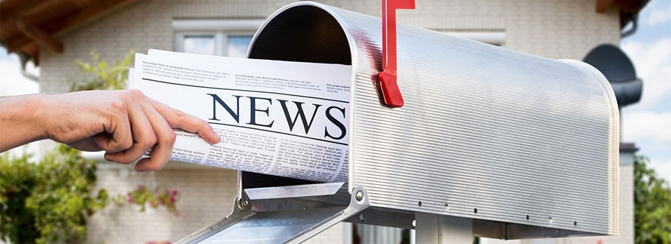 A person is removing a newspaper from a post-style mailbox. The red flag on the side of the silver mailbox is raised, indicating an item inside.