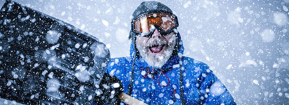 Man decked out in winter gear with a big smile with snow everywhere and holding a snow shovel