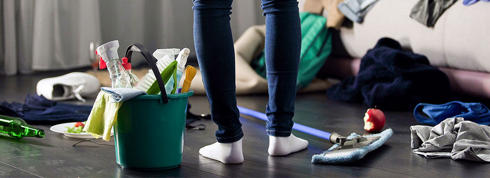 Woman standing in the middle of a messy bedroom with a bucket of cleaning supplies by her feet
