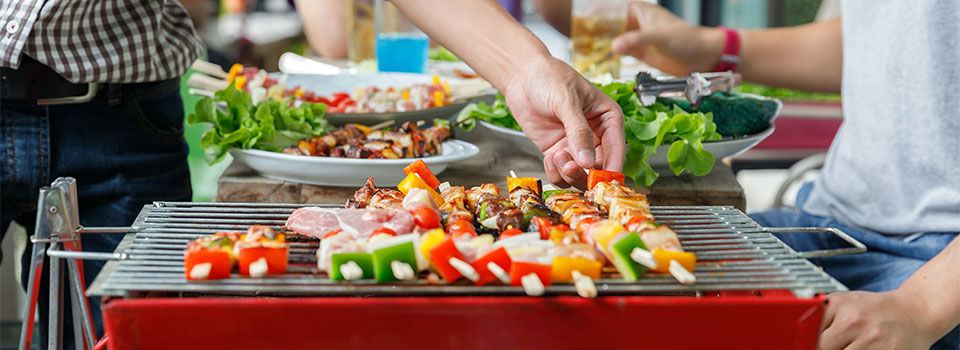 Vegetables kabobs grilling on a red grill with two people flipping them while they cook