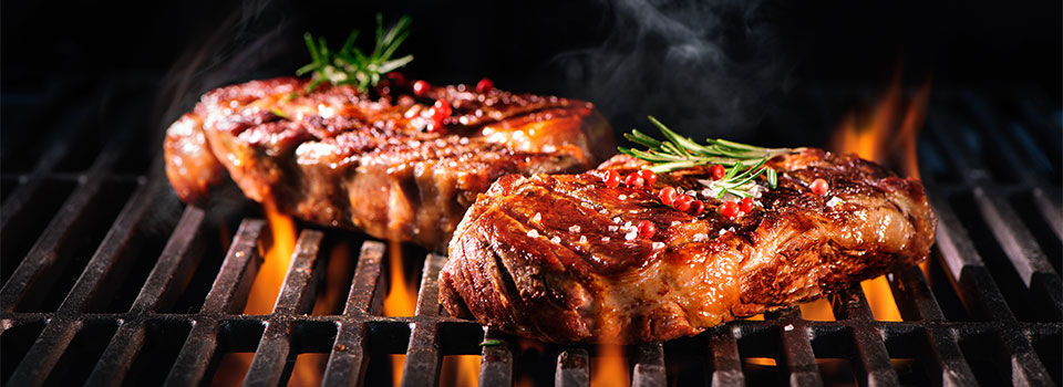 Two beautifully grilled steaks on an open flame, with sear marks and grill lines visible on the surface. The steaks are topped with herbs, which add a pop of color to the image. This appetizing and visually appealing image is sure to make your mouth water and inspire you to grill up some delicious steaks of your own.