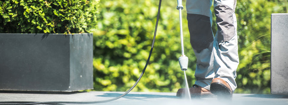 A man pressure washing a stone patio wearing gray pants and brown shoes. The patio has two stone planters with green bushes and a blurred out green background