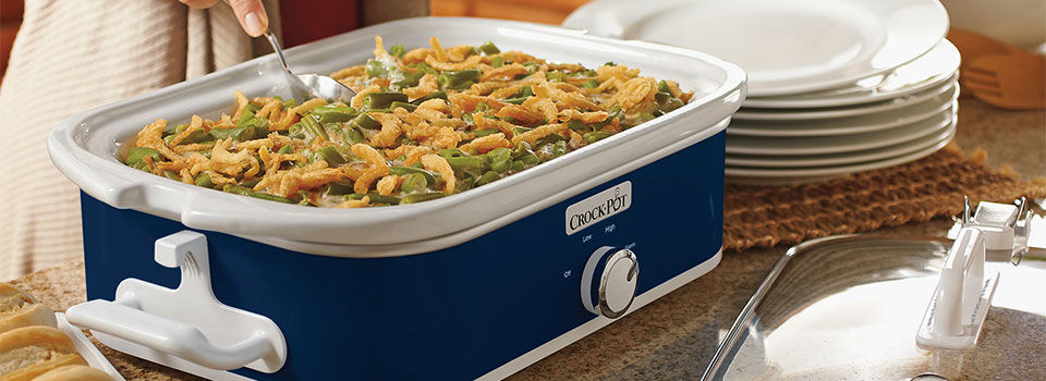 A blue and white slow cooker with green bean casserole inside. There is a woman spooning out a handful onto a white plate