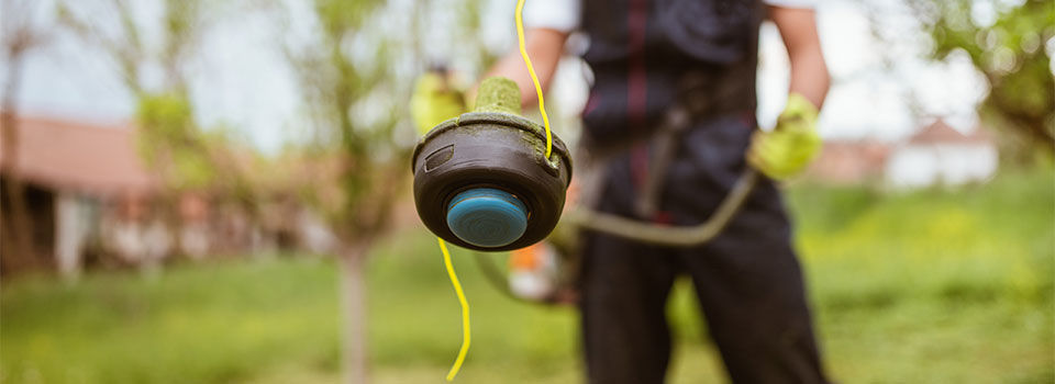 A close up image of a professional landscaper holding a string trimmer. The head of the string trimmer is in focus and the background is blurry