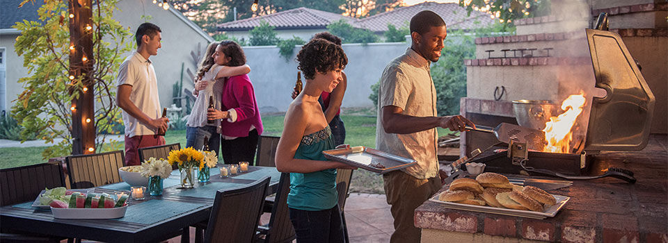 Multi ethnic hipsters enjoying backyard summer barbecue. The guests are greeting each other with hugs as they arrive. The African American male is manning the BBQ. Table is set with flowers, chips, watermelon and placemats.