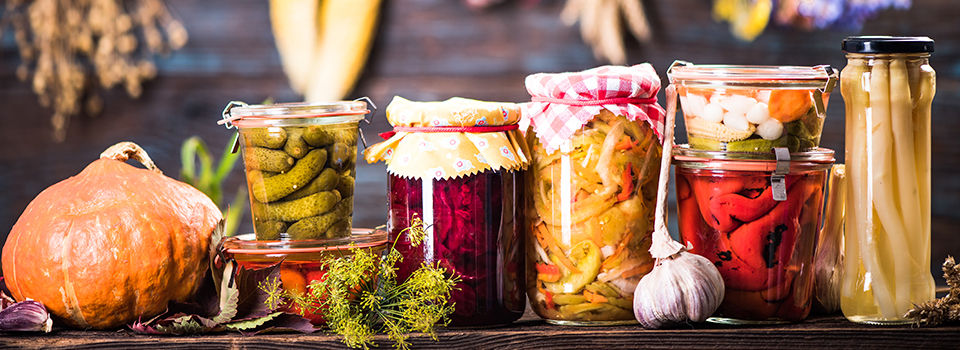 An assortment of fall vegetables in canning jars sitting on a dark wooden table surrounded by fall vegetables