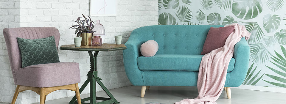 Pink chair and blue sofa with tropical leaf wallpaper and a white brick wall in the background