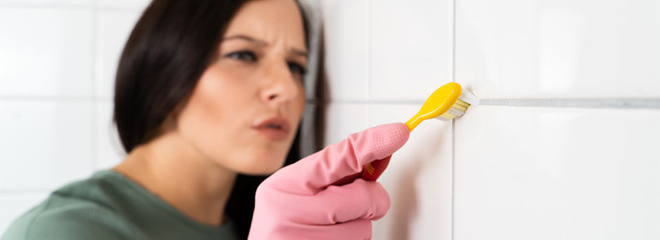 Woman cleaning bathroom tile grout using a yellow toothbrush while wearing pink rubber gloves