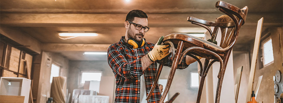 A young man wearing a flannel shirt, ear protection and work gloves, examining a wooden chair in his workshop