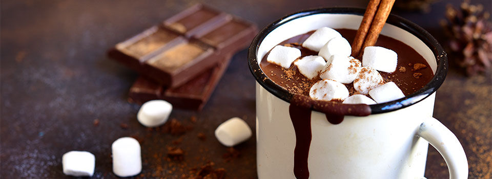 Cup of hot chocolate marshmallows and a cinnamon stick with chunk of chocolate in the background