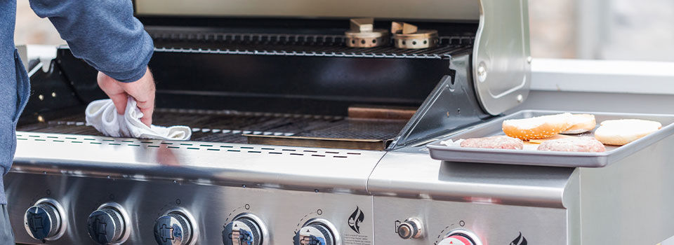 A person in a blue shirt uses a white rag to wipe off the grates of a stainless-steel backyard grill. The gas grill has adjustable knobs on the front. Two burgers and buns rest on a tray to the side of the grill.