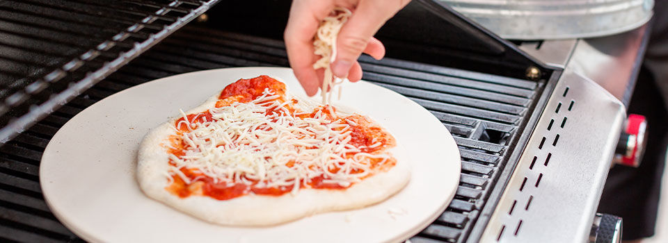 A person sprinkles cheese on a small pizza atop a tan cooking stone on a grill.