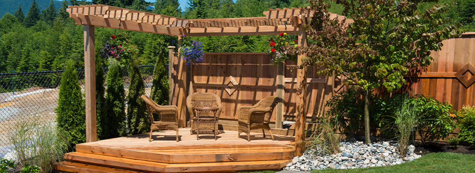 A landscaped yard hosts a cedar gazebo with three matching wooden chairs arranged in a circular formation. A wooden fence in the background has a similar cedar finish. 