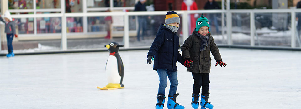 two young kids wearing winter clothing going ice skating. There is an inflatable penguin in the background.  