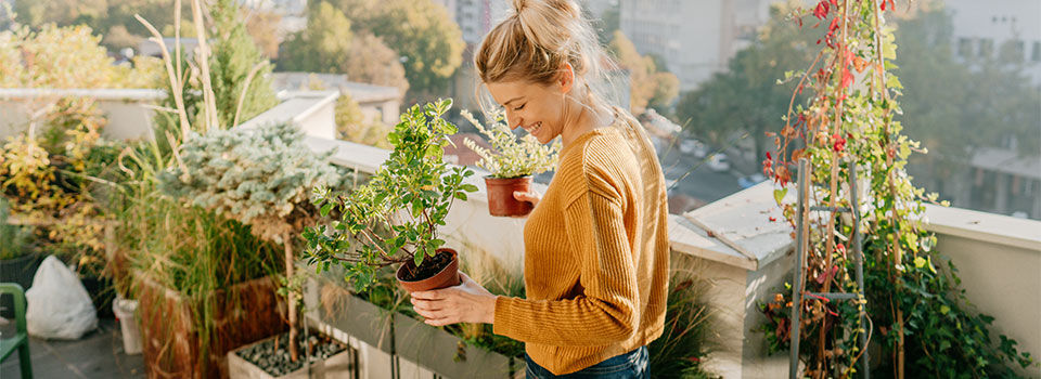 Woman on her balcony smile holding a potted plant in each hand surrounded by more plants