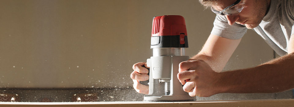 A person in a white shirt wearing safety goggles is using both hands to hold a silver and red wood router. Saw dust comes up as the person makes cuts using the router.