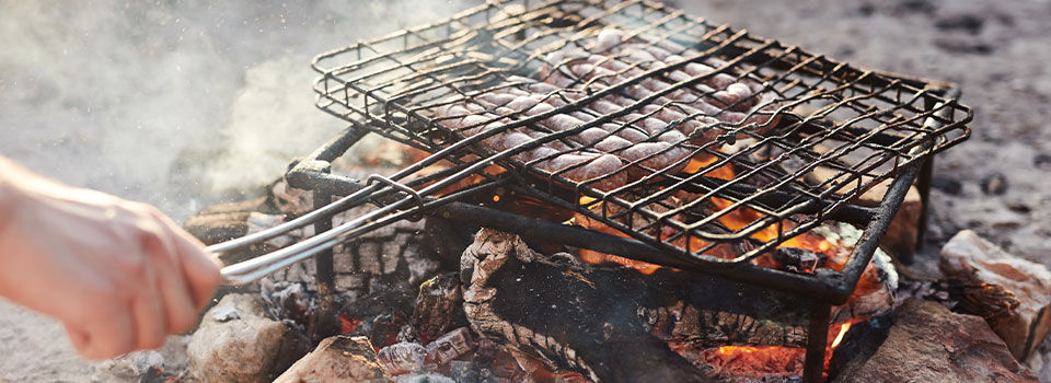 A person is holding the handle of a grilling basket, cooking sausages over a low fire and smoldering hot coals.