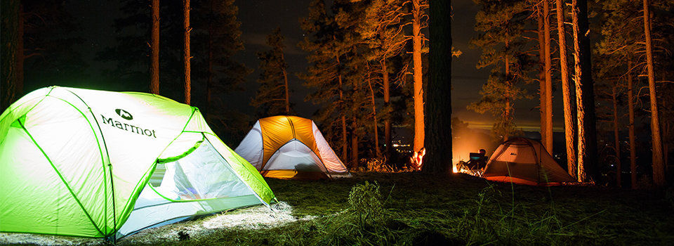 A bright green tent is shown in the center of a forest. The tent is lit from within, with several smaller orange tents in the background. 