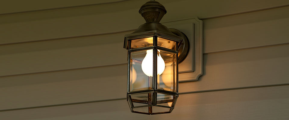 An outdoor light fixture is attached to a wooden wall. The light fixture is turned on and has a metal frame with glass panes.