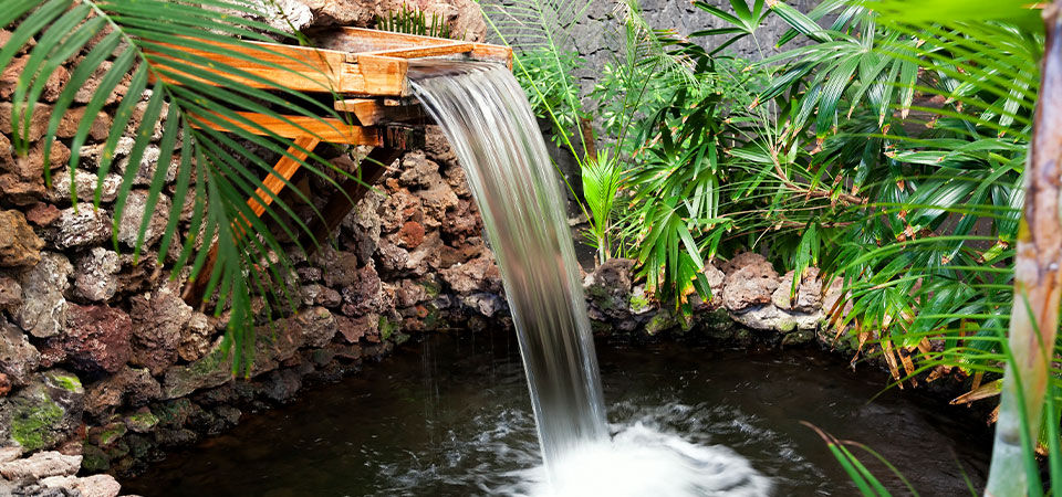 A tropical-themed backyard fountain and pond. The centerpiece is a large fountain made of wooden planks, with water running down its sides into the pond below. The pond is surrounded by rocks and tropical palm plants, adding to the overall aesthetic.