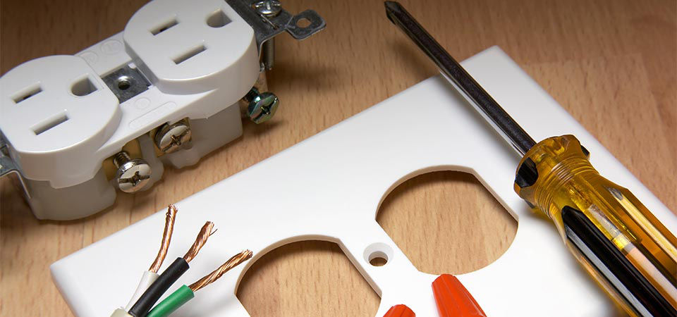 A white electrical outlet and the cover is shown on a wooden tabletop. There is a screwdriver and wiring on top of the outlet faceplate.