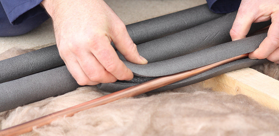 A close-up of a man putting insulation around pipes in an attic