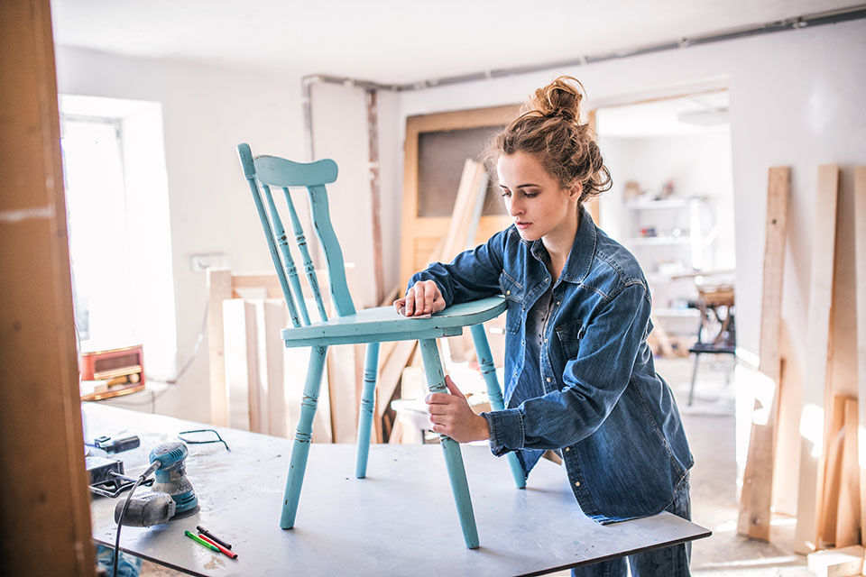 A woman wearing a denim jacket with her hair in a messy bun stands in the center of a bright white room, with woodworking projects in the background. She is sanding a turquoise-colored chair that has been painted with chalk paint.