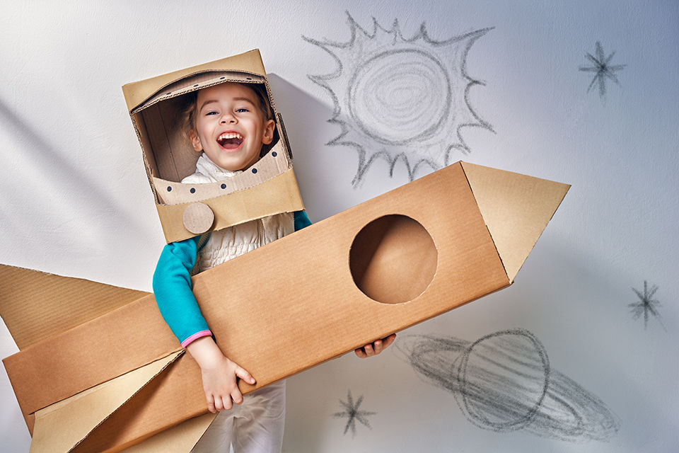 a little boy wearing an astronaut costume playing with a cardboard cut out rocket wearing a cardboard cutout astronaut helmet. There are drawings of a planet, stars, and sun on the wall behind him. 