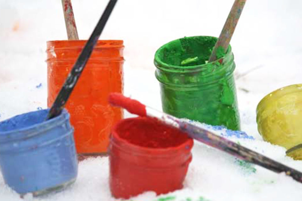 Five different color paint mason jars with paint brushes sticking out of the containers. They are sitting out side in the snow ready to paint the snow. 
