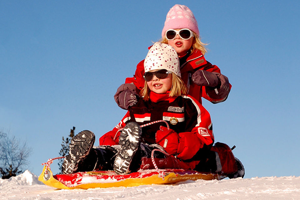 Two young girls going sledding on a bright winter day. They are wearing sunglasses and going down a hill on a tandem sled.