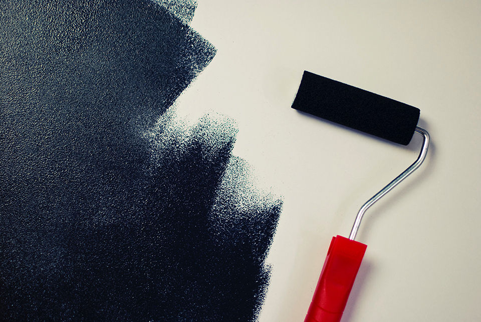 A red-handled paint roller is lying on a white surface that has been half-painted with black chalkboard paint. There is black chalkboard paint on the roller. 