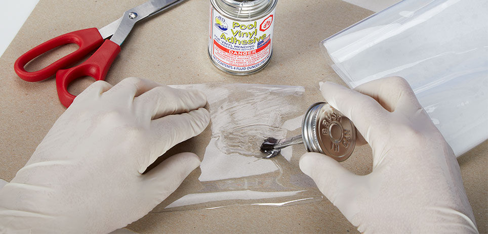 A person wearing gloves uses a brush to apply adhesive to vinyl for a pool liner repair. A small can of pool liner repair epoxy is on the surface near their hands, and a set of red-handled scissors is in the background.