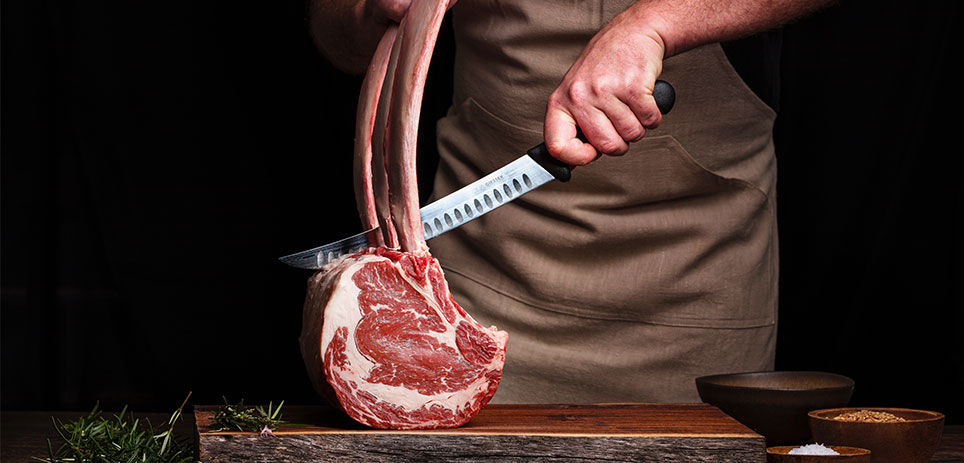 Close-up of a chef's hands holding a raw Tomahawk beef steak and a meat fork over a wooden chopping board. The steak is large and thick, with visible marbling and a bone-in shape reminiscent of a tomahawk. 