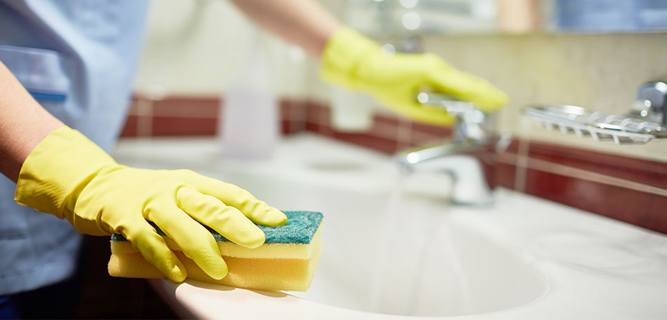 Person wearing yellow rubber cleaning gloves with a scrubber sponge in one hand while turning on bathroom sink faucet