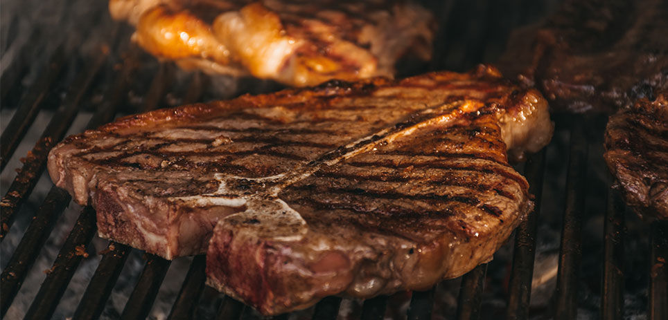 T-bone steak grilling on a barbecue grill. The steak is sizzling on the grates, with grill marks visible on the surface. The T-bone cut is evident, with a bone dividing the tenderloin and the strip steak, both of which are searing to a golden brown. The steak is generously seasoned with herbs and spices, adding to its aroma and flavor. 