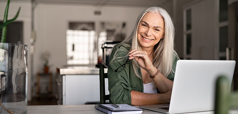 Woman smiling and holding her glasses sitting at a table with her laptop open