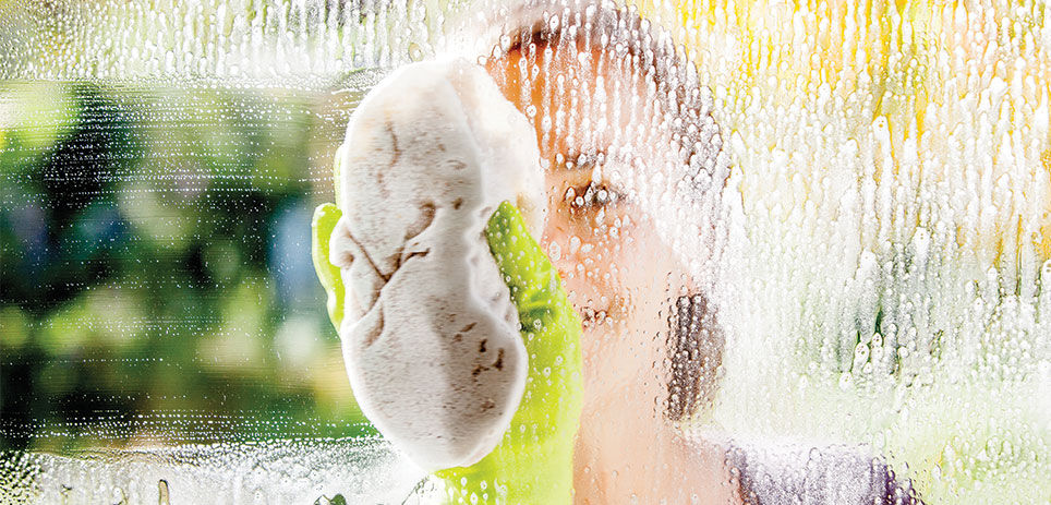 Woman wearing green rubber cleaning gloves on otherside of soapy window wiping it clean with a sponge