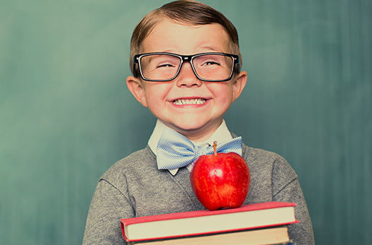 A young boy wearing thick rimmed glasses and holding a stack of books leans up against a chalkboard that reads back to school