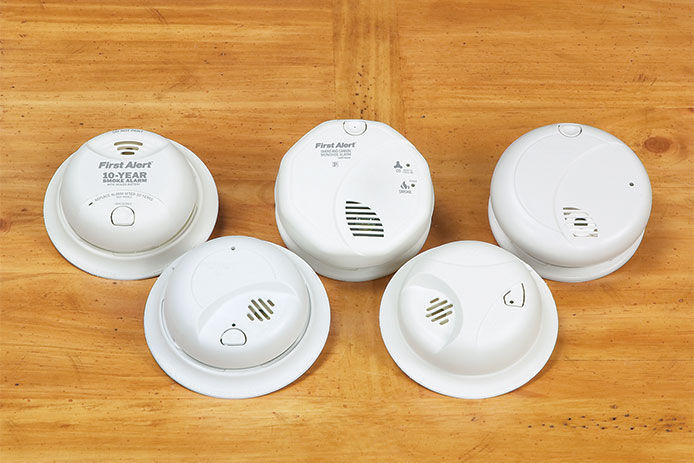 An image depicts five smoke alarms/carbon monoxide alarms arranged in a line on a wooden floor. Each alarm is small and white with a circular shape. The alarms have a test button and a red or green light to indicate the alarm status.