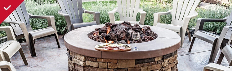 A circular stone fire pit filled with lava rocks and active flames is surrounded by several plastic Adirondack chairs on a patio, with a charcuterie board and wine glass resting on the ledge.