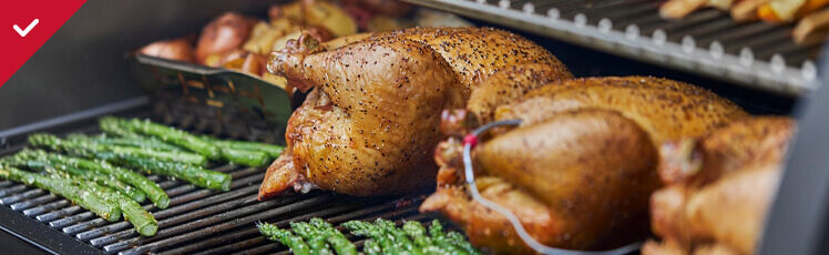 Two whole seasoned chickens, stalks of green asparagus, and halved red potatoes cook together on the metal grates of an outdoor grill or smoker.