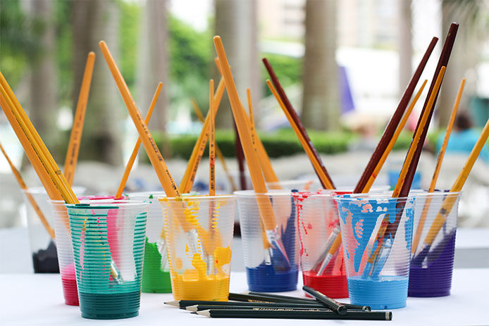 1 plastic cups filled with brightly colored children's acrylic paint and paint brushes in each cup. The cups are arranged on a white background and are evenly spaced. The paint colors include red, blue, green, yellow, purple, pink, orange, and black. The image is brightly lit, showcasing the vivid colors of the paint. The paint brushes are visible in each cup, with the bristles standing upright.