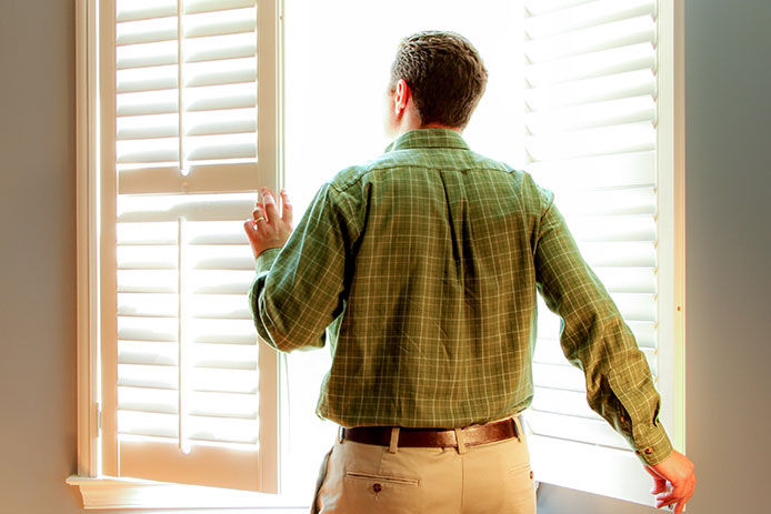 A man wearing a green flannel shirt, khaki pants, and a brown belt is seen in front of a partially open window. The window has wooden blinds on one side and a partially opened pane on the other side.