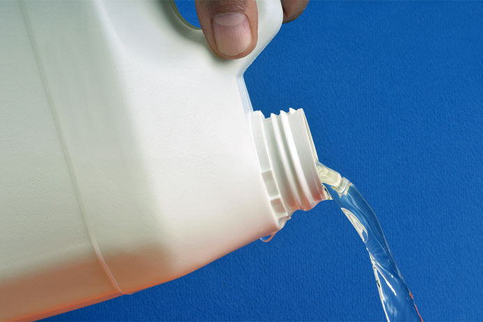 A gallon-size white chemical bottle being poured out of the bottle. There is a blue background. The chemical liquid appears to be flowing smoothly out of the bottle. The image depicts the use of industrial-strength cleaning chemicals and highlights the potential hazards associated with their use.